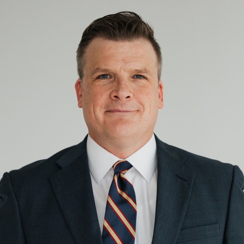 Professional headshot of a man in a navy suit, white shirt, and striped tie looking at the camera.