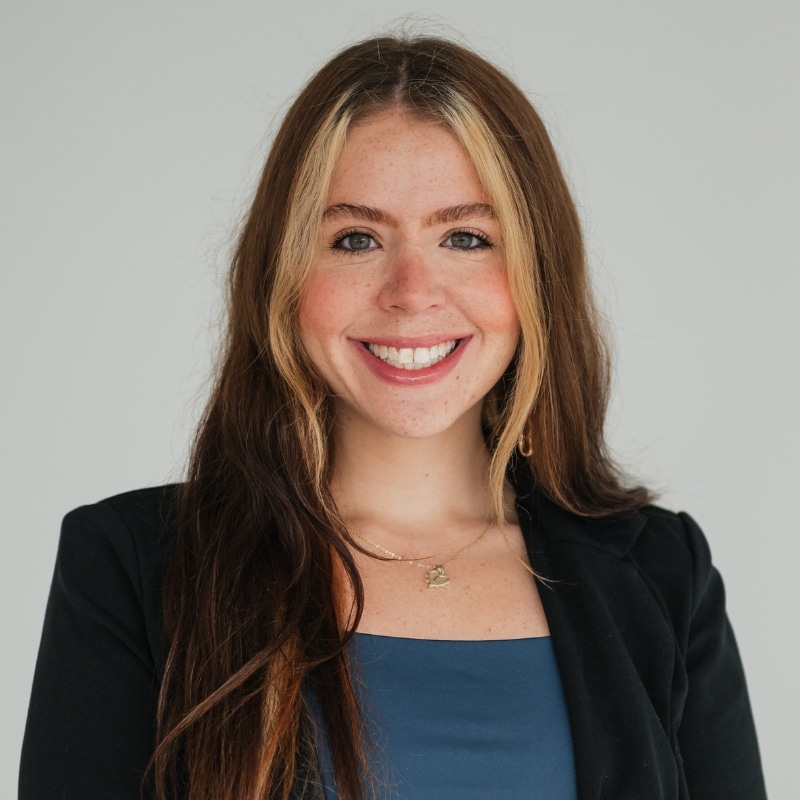 Smiling young woman with long brown hair and blonde highlights, wearing a black blazer and blue top, against a light gray background.