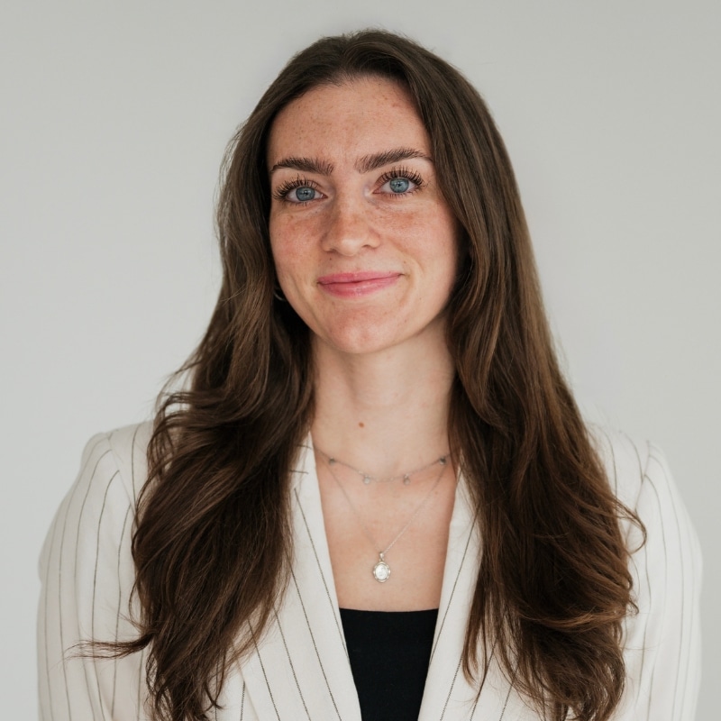 Portrait of a smiling woman with long brown hair, wearing a white pinstriped blazer and a pendant necklace.