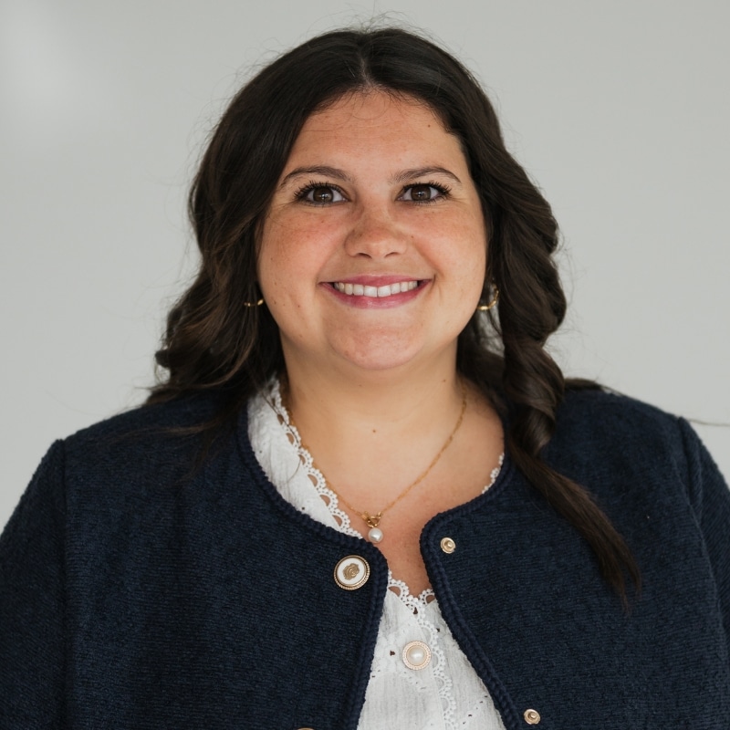 Smiling woman with dark hair in a navy blazer and white blouse, looking at the camera in a neutral background.
