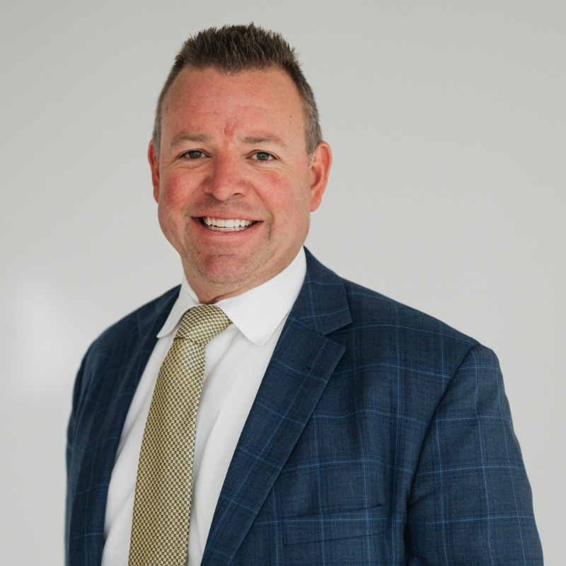 Smiling man in a blue plaid suit and beige tie posing for a business portrait against a light background.